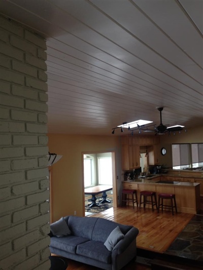 An open-concept living area with a white plank ceiling, light brick accent wall, blue sofa, and a kitchen with wooden cabinets and bar stools in the background.