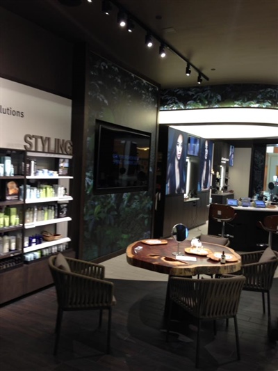 A stylish salon interior with product shelves labeled "STYLIST," a round table with chairs, and large mirrors with illuminated frames.