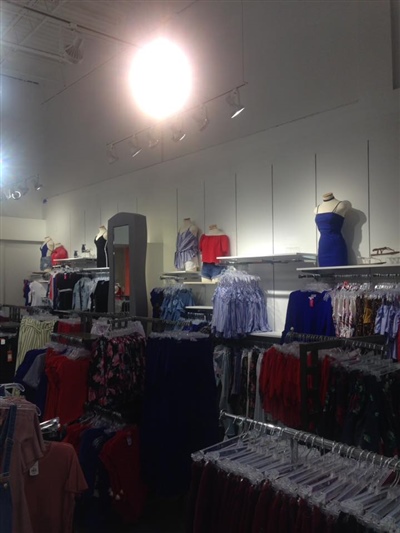 A clothing store with neatly arranged racks displaying various garments, mannequins on top shelves, and bright overhead lighting.