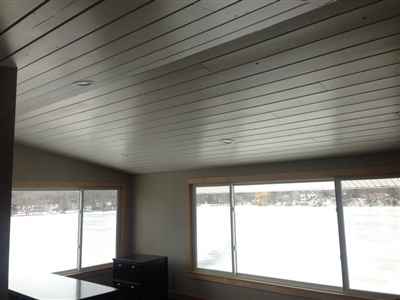 A room with a white plank ceiling, large windows offering a view of a snowy landscape, and minimal furnishings.