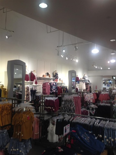 A clothing store interior with neatly arranged racks displaying various apparel, bright overhead lighting, and shelves with folded clothes and mannequins.