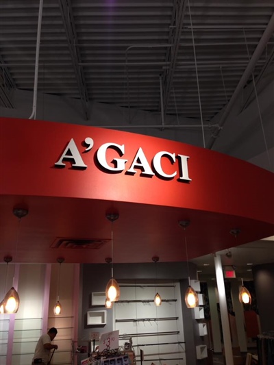 A red A'GACI store sign with bold white letters, hanging above the entrance in a mall, with the store interior visible below.