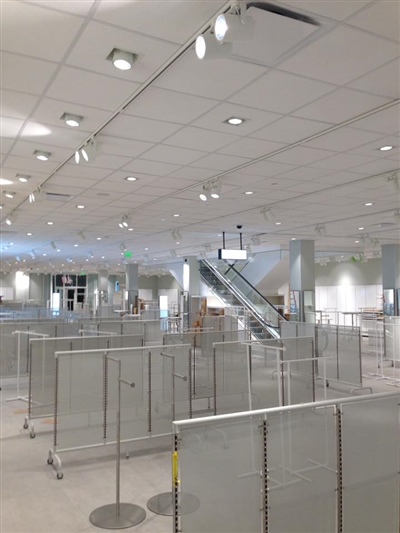 An empty retail store interior with numerous display racks, bright overhead lighting, and an escalator in the background.