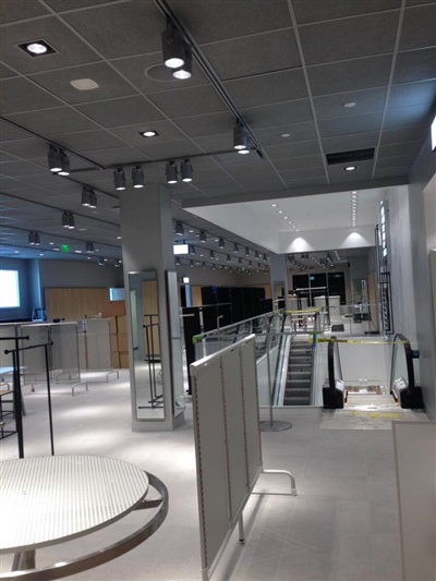 An empty retail store with modern lighting, various display racks, and an escalator in the background.