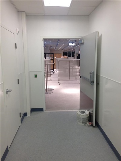 A corridor with white walls and gray flooring, leading to an open door that reveals an empty retail store area with display racks.