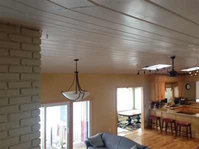 An open-concept living area with a white plank ceiling, light brick accent wall, hanging light fixtures, a blue sofa, and a kitchen with wooden cabinets and bar stools in the background.