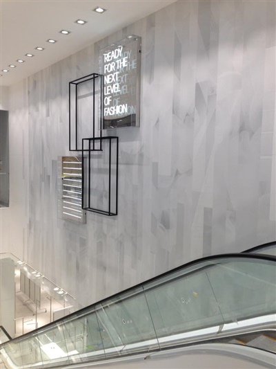 An escalator beside a white wall with modern geometric metal artwork and ceiling lights above.