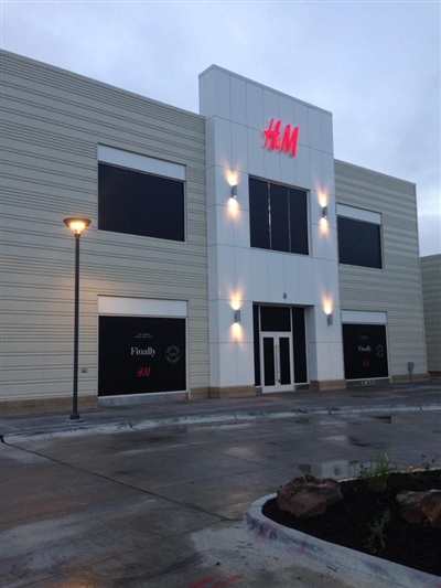 The exterior of an H&M store with a modern design, featuring large windows and illuminated signage, in a wet parking lot.