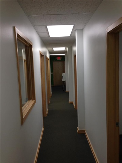 A narrow hallway with white walls, wooden door frames, and bright ceiling lights, leading to an exit door at the end.