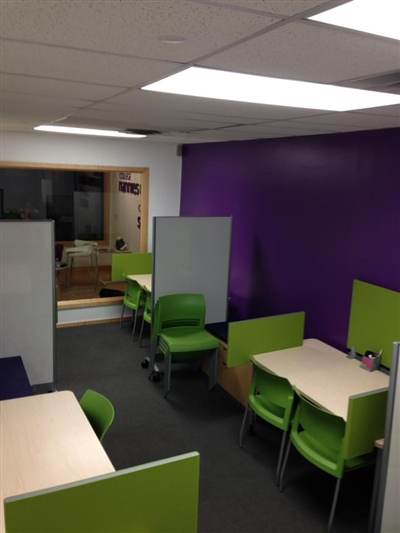 A small office area with green chairs, white desks, and purple accent walls, lit by ceiling lights.