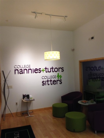 A welcoming reception area with a wall sign reading "College Nannies + Tutors + Sitters," furnished with green and purple seating, and illuminated by a hanging light.