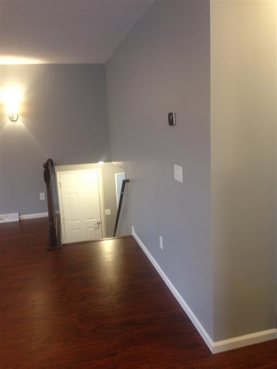 A room with gray walls, dark wood flooring, and a staircase leading down, illuminated by a wall sconce.