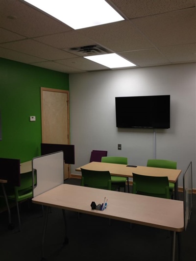 A small meeting room with green accent walls, green chairs, desks, and a wall-mounted TV, lit by ceiling lights.