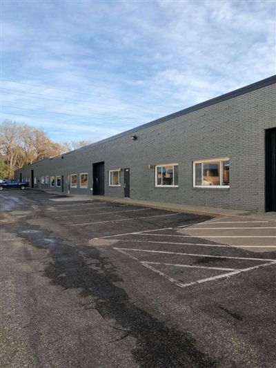 A long, single-story industrial building with several windows and doors, adjacent to a parking lot under a clear sky.