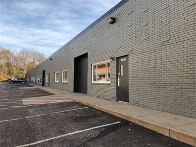 A long, gray brick industrial building with windows and a door, next to an empty parking lot under a clear sky.