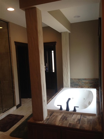 A bathroom with beige walls, a large built-in bathtub with dark fixtures, wooden accents, and an open doorway leading to a hallway.