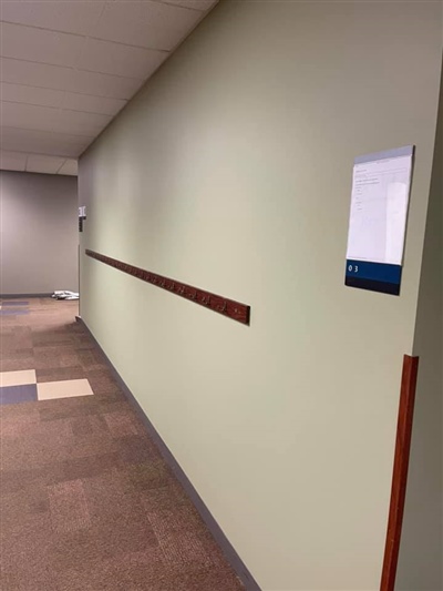 A hallway with cream-colored walls, a wooden chair rail, and a wall-mounted sign, with carpeted flooring.