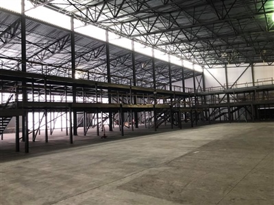 A spacious, empty industrial warehouse with high ceilings, metal beams, and a multi-level structure, illuminated by natural light through the roof.