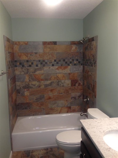 A bathroom with a bathtub and shower featuring a natural stone tile wall, a toilet, and a sink with a marble countertop.