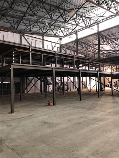 An empty industrial warehouse with metal beams, high ceilings, and a mezzanine level, with a traffic cone on the concrete floor.