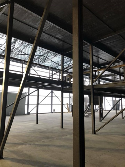 An interior view of an empty industrial warehouse with metal beams and a high ceiling, featuring a mezzanine level.