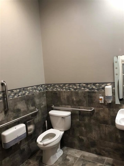 A clean, modern restroom with a toilet, grab bars, a sink, and tiled walls.