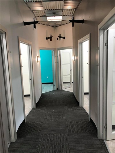 A hallway with white walls and doors, carpeted flooring, and ceiling lights, leading to rooms with one door open showing a blue wall inside.