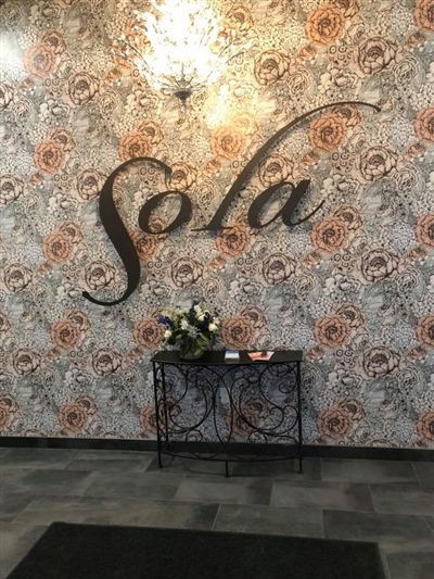 A decorated wall with the word "Sola" in cursive, above a small table with flowers, against a patterned wallpaper background.