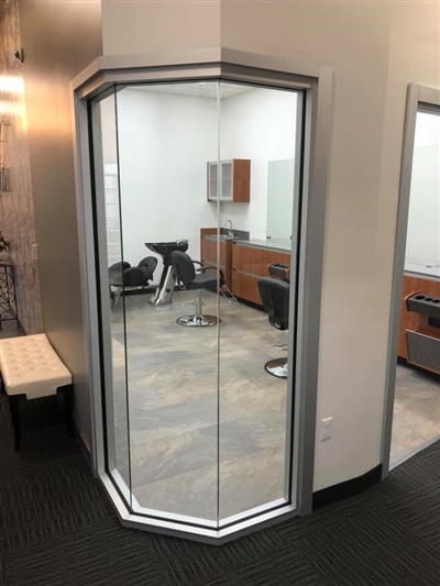 A modern hair salon room with glass walls, featuring a chair, mirrors, and wooden cabinets inside.