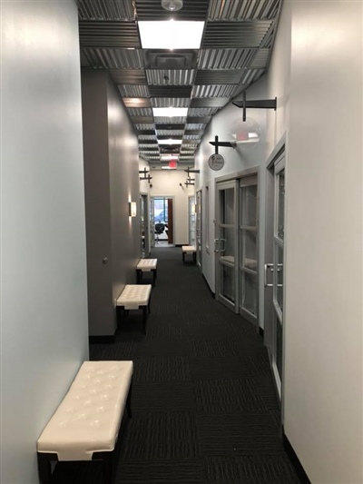 A long hallway with white walls, carpeted flooring, ceiling lights, and benches along the sides, leading to glass doors at the end.