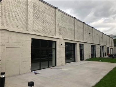 The exterior of a modern industrial building with large black-framed windows and doors, adjacent to a concrete patio and a grassy area.