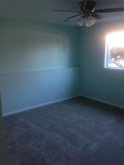 An empty room with light blue walls, a gray carpet, a ceiling fan, and a window letting in natural light.