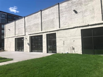 The exterior of a modern industrial building with large black-framed windows and doors, a concrete patio, and a grassy lawn under a clear blue sky.