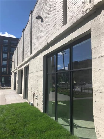 The exterior of a modern industrial building with large glass doors and windows, adjacent to a grassy area and concrete patio, under a clear blue sky.