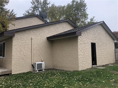 The exterior of a beige residential house with a sloped roof, an air conditioning unit, and a grassy yard, surrounded by trees.