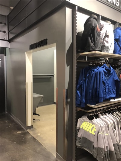 An open door leading to a fitting room area in a retail store, with clothing racks displaying Nike apparel nearby.