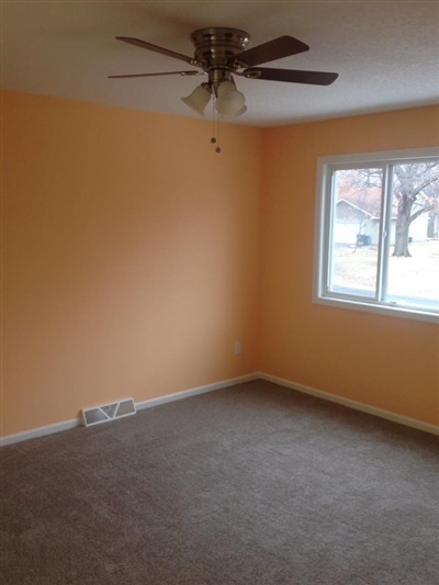 An empty room with orange walls, a gray carpet, a ceiling fan, and a window showing a view of the outside.