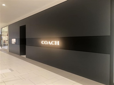 A sleek, modern exterior of a Coach store with a black facade and illuminated Coach logo, located in a shopping mall.