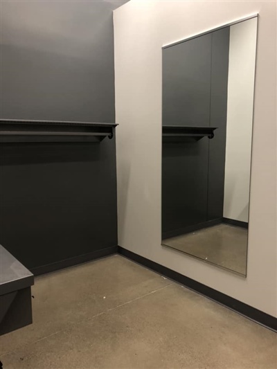 A fitting room with gray walls, a large mirror, and a bench on a concrete floor.