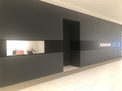 A sleek, modern exterior of a Coach store with a black facade, an illuminated Coach logo, and a display window showcasing products, located in a shopping mall.