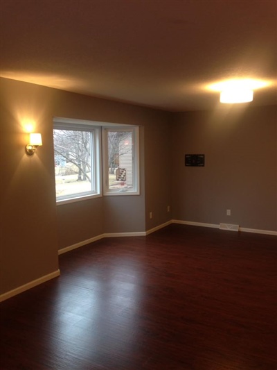 An empty room with taupe walls, dark hardwood floors, two windows, a wall sconce, and a ceiling light.