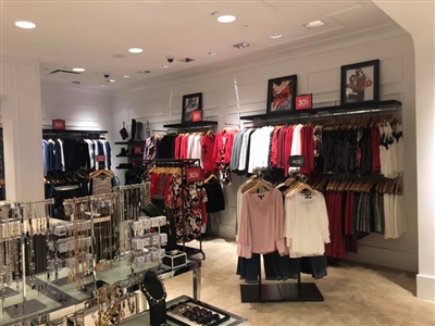 A retail store interior with neatly arranged clothing racks displaying women's apparel, accessories on glass shelves, and framed promotional images on the walls.