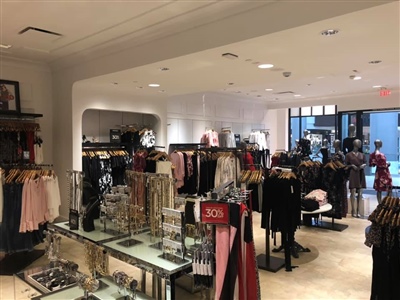 A retail store interior featuring neatly arranged clothing racks with women's apparel, glass display shelves with accessories, and well-lit, stylish decor.