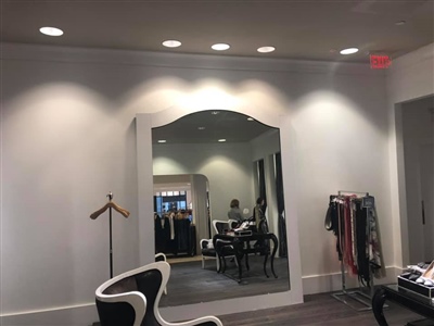 A fitting room area in a retail store with a large decorative mirror, elegant seating, and clothing racks, illuminated by recessed ceiling lights.