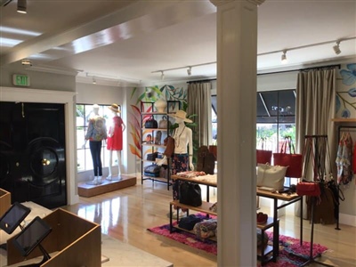 A boutique store interior featuring mannequins in the display window, various clothing racks, a table with accessories, and a cozy, well-lit atmosphere.