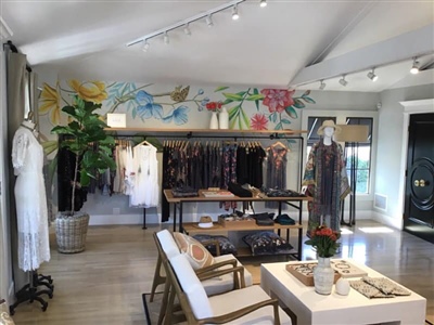 A boutique store interior featuring a colorful mural on the wall, clothing racks with a variety of apparel, a table with accessories, and comfortable seating in a bright, well-lit space.