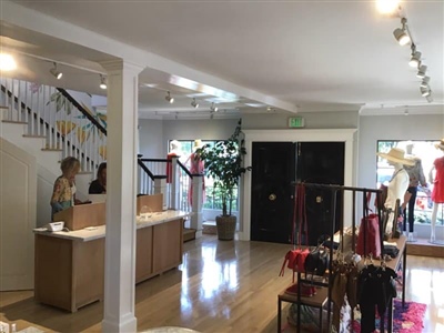 A boutique store interior featuring a checkout counter, clothing racks with various garments, mannequins in the display window, and a staircase, all in a bright, welcoming atmosphere.