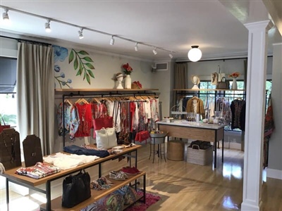 A boutique store interior featuring neatly arranged clothing racks with colorful garments, tables displaying accessories, and a bright, inviting atmosphere with decorative touches.