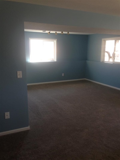 An empty room with blue walls, a gray carpet, and two windows letting in natural light.