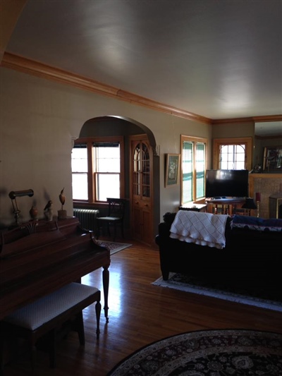 A living room with a wooden piano, a black couch, a TV, hardwood floors, and large windows letting in natural light.